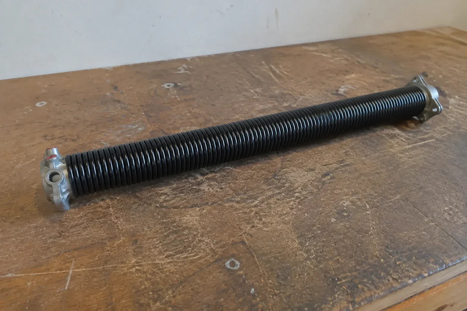 Detached black coil spring with metal end fittings on a workbench. — Torsion Spring Replacement, Markham