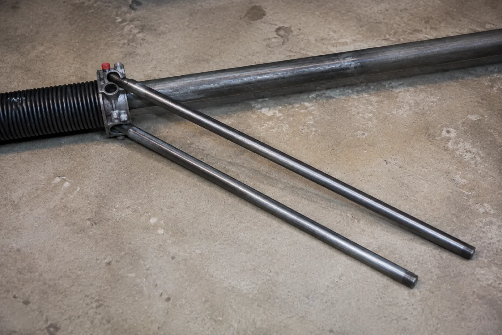Two long steel winding bars inserted into the cone of a black torsion spring lying on a concrete floor.